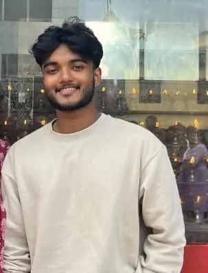 Smiling young man in a beige sweatshirt standing in front of a glass wall with warm lights in the background – freelance digital marketing expert in Calicut."